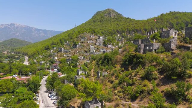 Kayakoy, Turkey. Hundreds of abandoned stone house ruins cascading down a forested mountainside, aerial drone view under clear blue sky with distant peaks. Aerial View