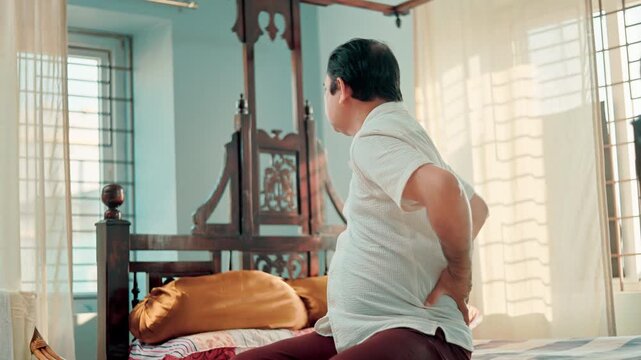Mature Indian male struggling with lower back pain while sitting on a bed at home.