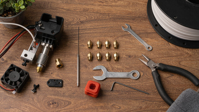 A wooden table displays tools and parts for a 3D printer. Components include a hotend assembly, nozzles, wrenches, and a spool of filament. Tools are neatly arranged for easy access