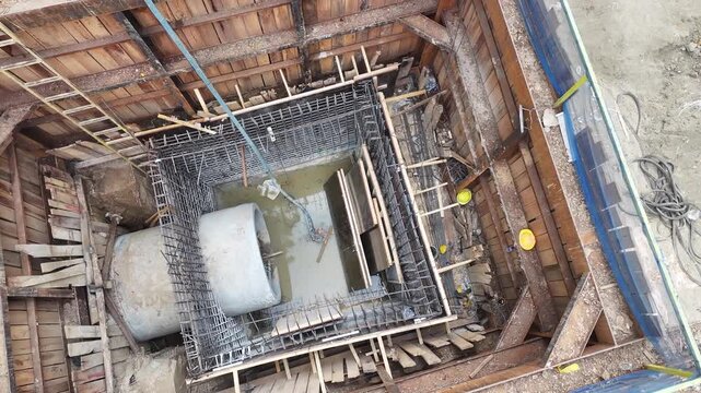 Workers build foundations near a calm river. Steel beams and wooden frames stand tall. Red pipes lie scattered on dirt ground. People move carefully through the construction site street construction