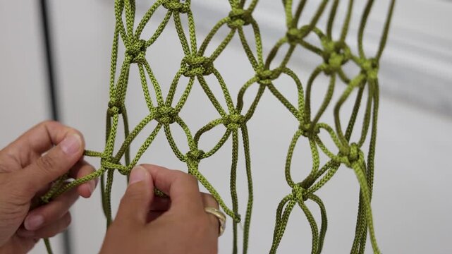 Close-up of hands creating a decorative green macrame piece using intricate knotting techniques, showcasing handmade craftsmanship, textile artistry and traditional fiber work.