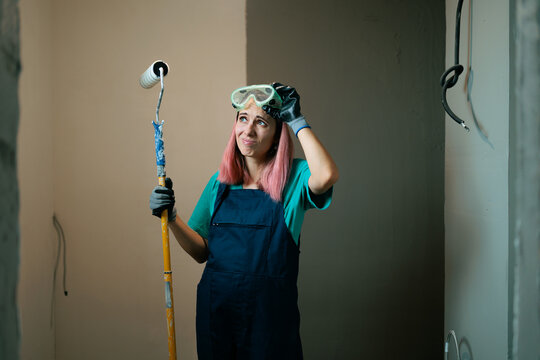 Painter Looking Confused Adjusting Safety Goggles Indoors. Clueless young woman worker holding paint roller tool touches safety goggles thoughtfully inside