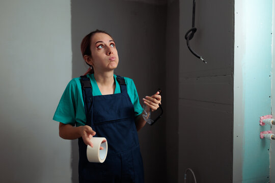 Pensive Female Worker Holding Masking Tape Looking Up Upset. Tired young woman painter holding adhesive tape holds protective glasses stands indoor thoughtfully