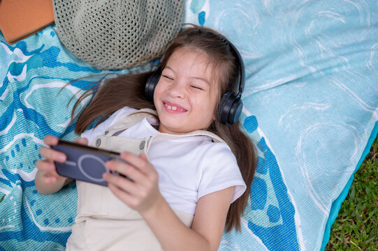 Young mixed race girl wearing headphones using smartphone while happily lying on a blanket in park