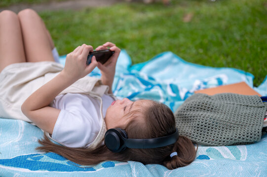 Young girl wearing headphones relaxes on a blanket while using her smartphone in the park.