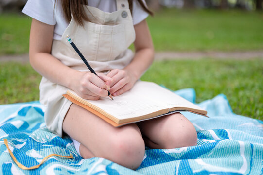 Close up view of a young girl kid writing with pencil in notebook on picnic blanket in the park.