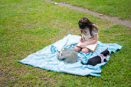 Young girl writing or drawing in notebook on a picnic blanket beside a dog in the park.