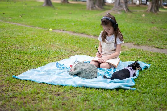 Young girl writing or drawing with pencil in notebook on blanket while relaxing with dog in the park