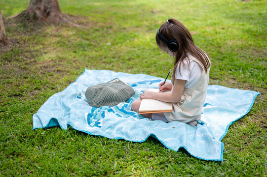 Side view of a young girl wearing headphones writes in a notebook while sitting on a blanket in park