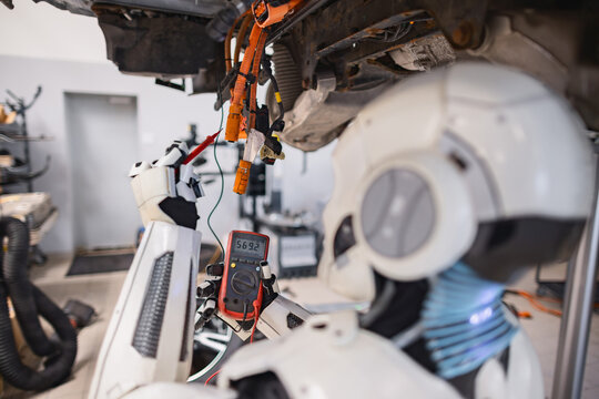 A humanoid robot uses a multimeter to test electrical wiring on a vehicle chassis in a workshop setting