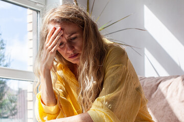 Woman suffering from headache and migraine holding forehead with hand sitting near window at home....