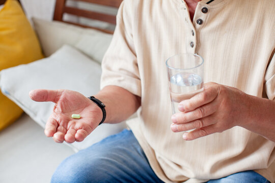 Senior man holding pill and glass of water in hand at home. Daily medication routine and prescription drug intake. Elderly healthcare and medical treatment for chronic disease concept
