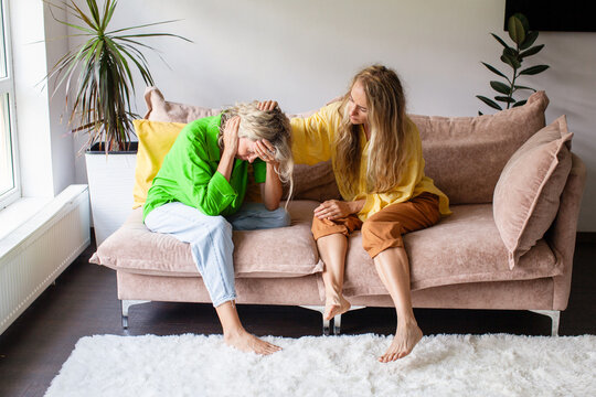 Woman crying on sofa while friend comforting her. Emotional support and mental health problem concept. Empathy, stress and depression help with anxiety and grief at home interior