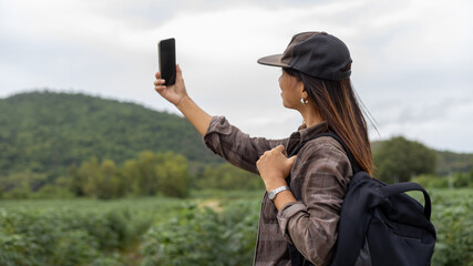 A woman stands in a green field with hills in the background. She holds her phone up to take a selfie. The sky is cloudy and she has a backpack on her shoulder © Happy Photo