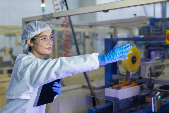 woman worker in manufacturing factory wearing safety hairnet and lab coat pointing at production equipment holding clipboard showing attentive professional expression during inspection
