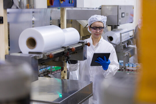 Caucasian woman worker in modern factory cleanroom wearing white uniform inspecting large plastic roll machine with digital tablet looking focused and productive during manufacturing process