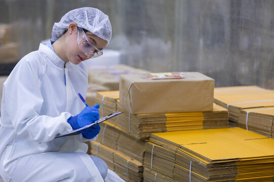 Female worker wearing protective uniform performs quality inspection writing on clipboard near stack of cardboard box in warehouse environment showing diligent attention to detail