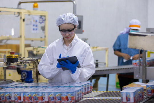 Female factory worker in white coat and hairnet doing inspection with digital tablet on manufacturing production line feeling focused and attentive to quality control process of packaged product