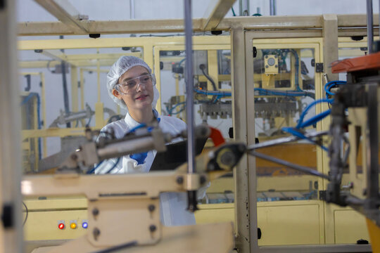 Content adult woman worker inside modern factory operating heavy machinery wearing protective safety eyewear and hygienic hairnet for manufacturing industry