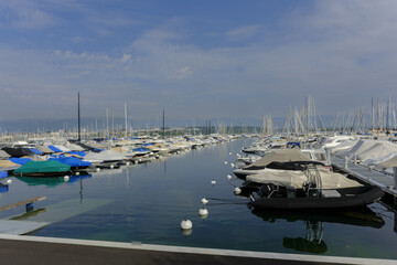 Fototapeta premium Covered with tarps, the yachts in the Geneva marina line the pier in soft light, their tall masts rising over the still lake. 