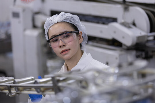 asian female worker wearing safety glasses and white hairnet checking manufacturing production line inside modern industrial factory facility with serious expression