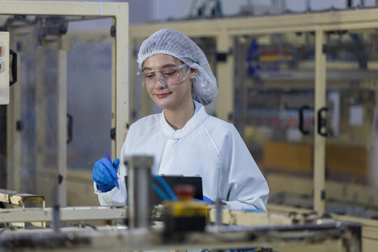 Confident female worker in white uniform and hairnet conducting safety inspection in manufacturing factory using digital tablet for modern production monitoring with focused professional smile