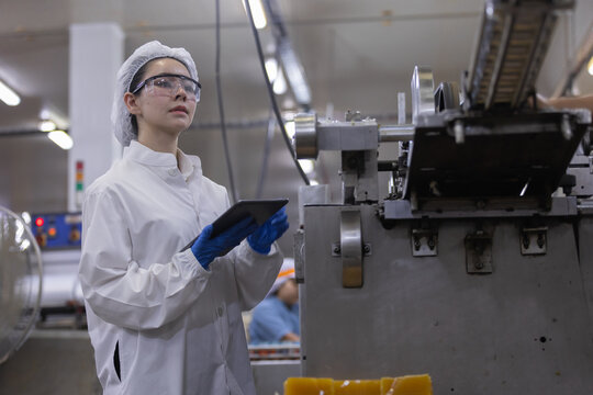 Asian female worker conducts inspection on production line machinery with digital tablet maintaining focused expression wearing protective gear inside modern manufacturing facility
