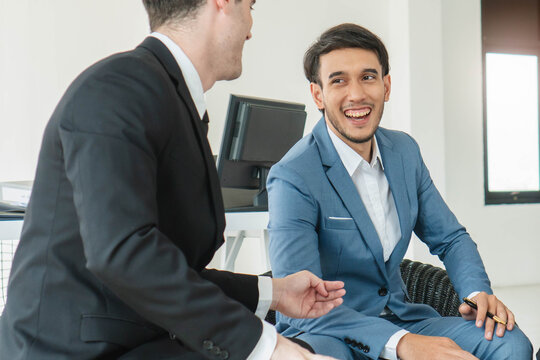 Two diverse professional businessmen in formal suits having a friendly discussion and laughing together during a successful corporate meeting in a modern office