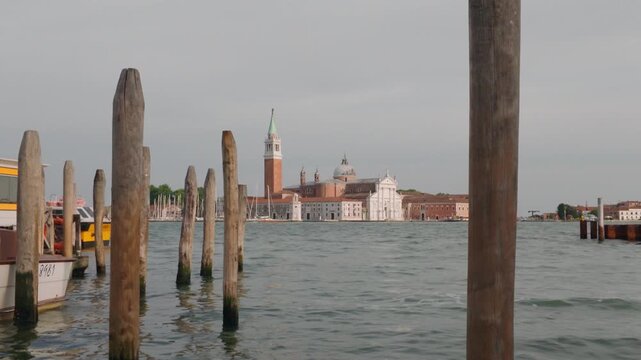 Scenic view across the Venetian Lagoon toward San Giorgio Maggiore, framed by wooden mooring poles and boats under an overcast sky in Venice, Italy.