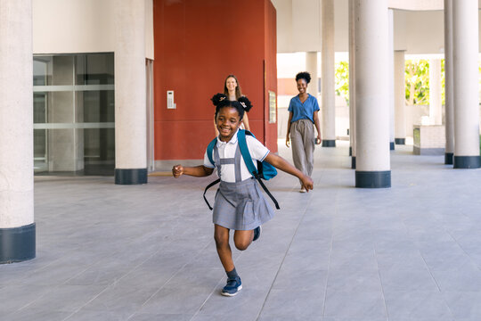 Happy diverse schoolgirl in uniform running on first day back to school, her mothers walking behind her