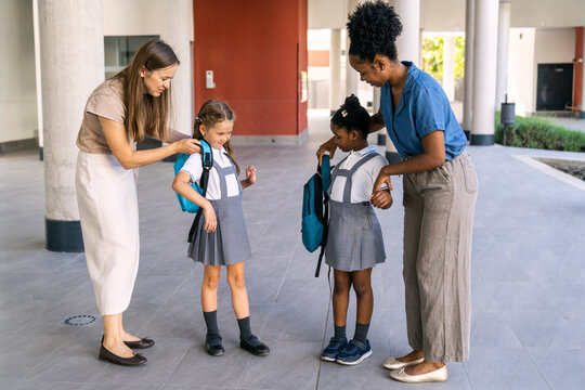 Multiethnic mothers assisting their young daughters, dressed in school uniforms, preparing for their first day back to school