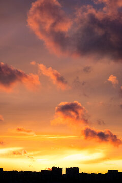 Vertical view of a dramatic orange sunset sky with dark clouds and silhouettes of residential buildings at the horizon. The warm golden and deep purple tones create a moody atmospheric evening scene.