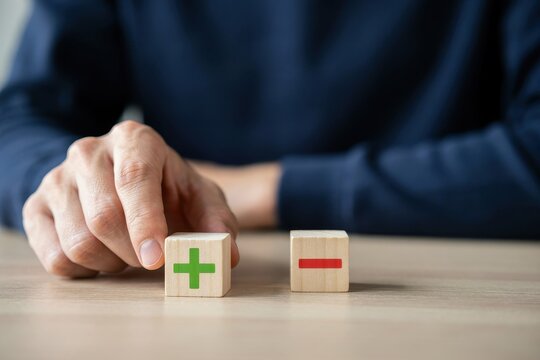 A person holding a wooden block with a green plus sign next to a minus sign on a table. This image is ideal for business decision-making and risk analysis concepts.