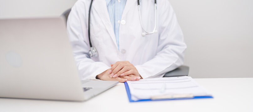 Professional doctor wearing lab coat and stethoscope using laptop computer and taking report notes on notebook at desk in hospital clinic office, medical technology and healthcare concept
