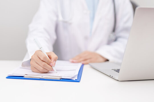 Professional doctor wearing lab coat and stethoscope using laptop computer and taking report notes on notebook at desk in hospital clinic office, medical technology and healthcare concept