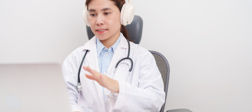 Professional doctor wearing headphone and lab coat using laptop for telehealth, telemedicine, online consultation and medical technology video meeting at desk in hospital office concept