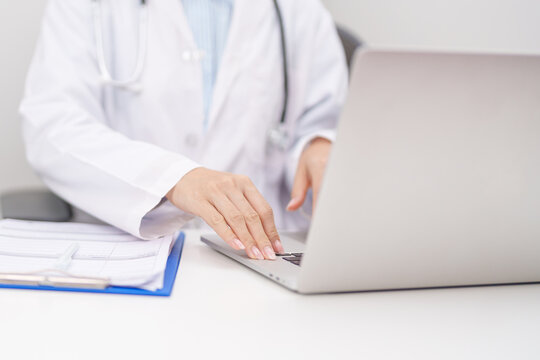 Professional doctor wearing lab coat and stethoscope using laptop computer and taking report notes on notebook at desk in hospital clinic office, medical technology and healthcare concept