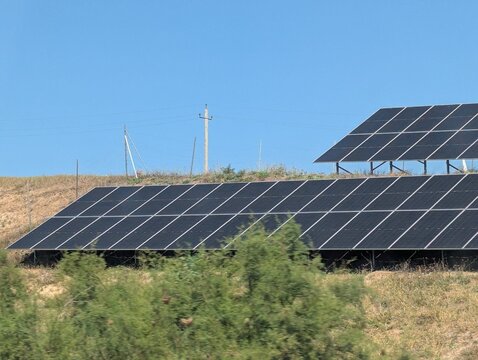 A sprawling field of solar panels stretches across a sun-drenched hillside in the Georgian countryside, soaking up the intense summer rays. The gleaming black and blue modules contrast 