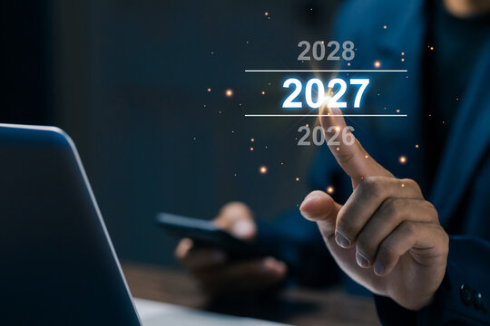 Business professional selects 2027 on a futuristic digital interface beside laptop, representing new year planning, future goals, business strategy, innovation, progress, and technology.