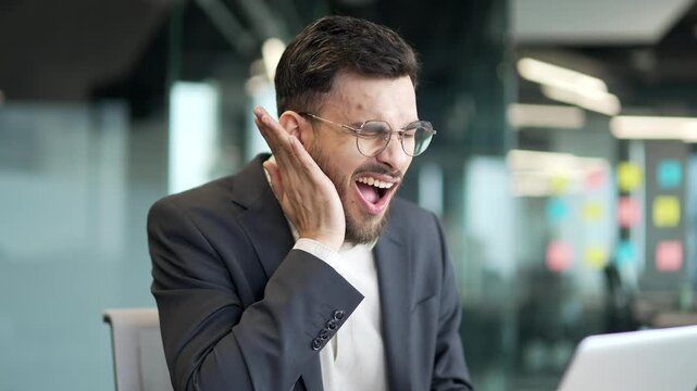 Upset businessman in formal suit suffering from toothache while sitting at workplace in business office. The worker holds his hand on his cheek, massaging sore spot. He needs to see dentist. Close up