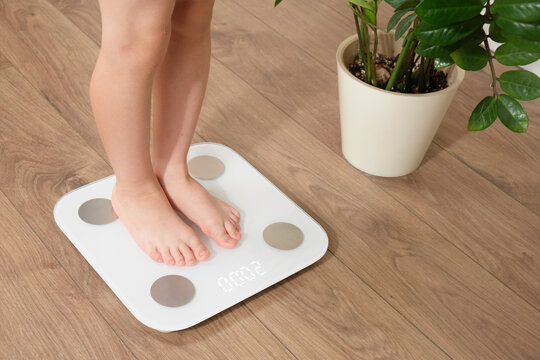 Young boy standing on digital bathroom scale at home. Child growth monitoring and healthy weight measurement concept with plant and wooden floor. Kid aged five years