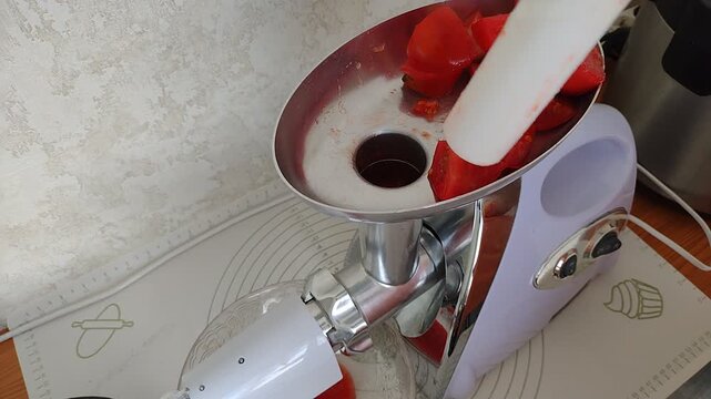 A hand pressing sliced red tomatoes with a plastic pusher into an electric juicer tray. stock footage for kitchen appliances, tomato sauce prep, food processing, and home canning vlogs.