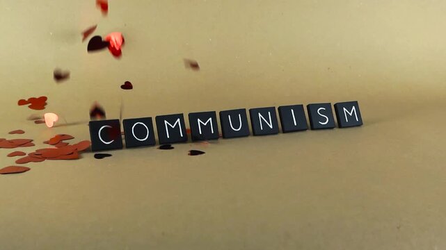 Red heart shaped confetti gently falling in slow motion over the word communism, spelled out with black tiles, creating a conceptual representation of political ideology and emotion