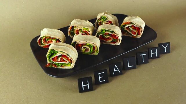 Delicious lavash pinwheel rolls filled with fresh vegetables and ham resting on a black plate with the word HEALTHY spelled out next to them, representing a concept of proper nutrition