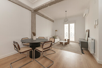 Fototapeta premium Living-dining room with exposed concrete beam, black round table, cane chairs, sofa, and balcony view