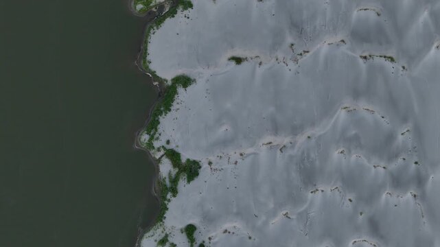 Low water level exposes broad sediment along the Po River during the 2026 drought in northern Italy, with sparse shoreline vegetation and shallow water, top down drone flyover.