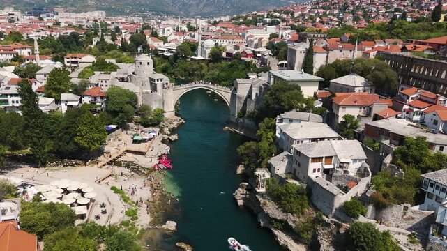 Historic Stari Most stone bridge spans the Neretva River, as a drone flies upstream over busy riverside beaches and the surrounding old town.