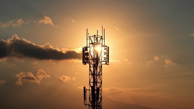 Cell phone signal tower silhouetted at sunset with clouds. Telecommunication, connectivity, and network infrastructure concept footage.