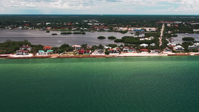 Upscale single-family beach houses line a thin sand strip beside bright teal coastal water as the drone glides parallel offshore.
