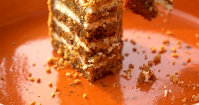 closeup a fork picking up a carrot cake bite beside the remaining slice with moist brown sponge pale cream layers loose nut crumbs and smears across a vivid orange plate